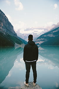 man in black hoodie and black pants standing in front of body of water and mountain during daytime thumbnail