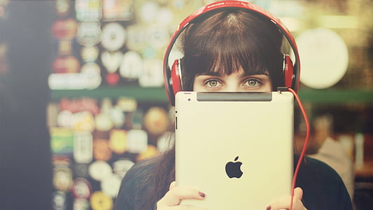 selective focus photography of woman wearing red corded headphones while holding silver iPad thumbnail