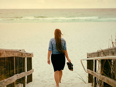 woman holding pair of sandals while walking on sand thumbnail