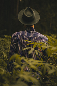 person wearing black cowboy hat and black and white dress shirt standing and surrounded by green plants thumbnail