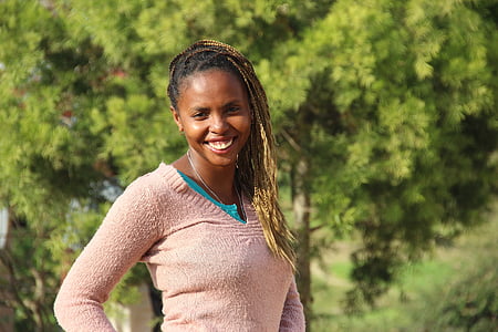 smiling woman in gray v-neck sweater standing near green trees thumbnail