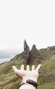 person's hand extending towards grey boulders surrounded by green grass under white cloudy daytime sky thumbnail