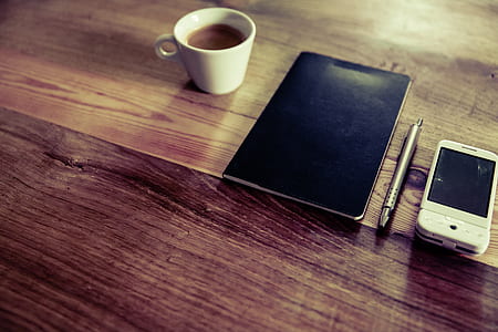 black notebook near click pen, white phone and white ceramic teacup with coffee thumbnail