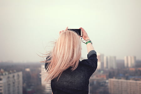 women wearing black denim jacket holding white smartphone while capturing a photo of city high rise buildings thumbnail