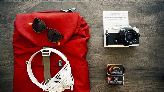 white full size corded headphones, sunglasses on red leather backpack beside gray SLR camera thumbnail