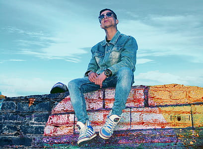 Man in Gray Denim Jacket and Jeans Sitting on Brick Wall thumbnail