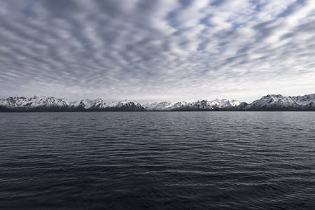 calm body of water in front of black snow capped mountain under gloomy sky thumbnail