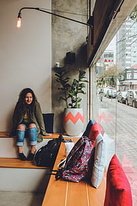 Woman in Gray Zip Up Jacket and Blue Distressed Jeans Sitting on Bench Near Backpack Looking Outside in Room thumbnail