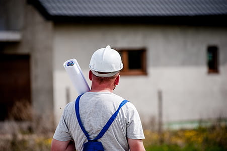 man wearing white hard hat, gray shirt and blue rompers carrying blueprints facing gray concrete building thumbnail
