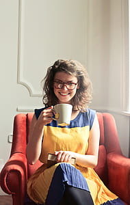 Young Woman Wearing Yellow And Blue Dress With Spectacles, And Holding A Book While Enjoying A Cup Of Tea At Home thumbnail