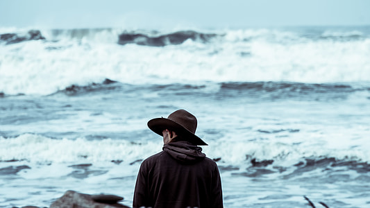 man standing while wearing hat in front of body of water with wave during daytime thumbnail