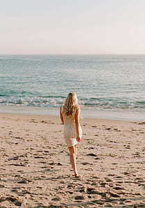 woman wearing white lace sleeveless dress walking on seashore near beach under cloudy skies thumbnail