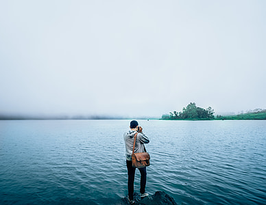 man wearing grey jacket and carrying bag standing on rock near body of water thumbnail