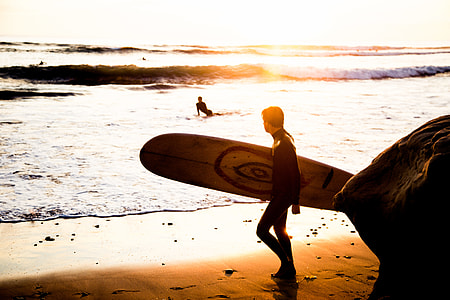 man walking on seashore holding surfboard during sunset thumbnail