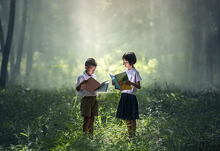 girl and boy holding book while standing in grass field thumbnail