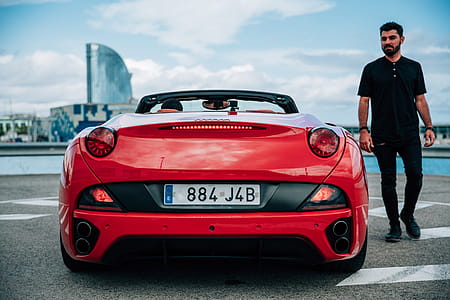 A bearded young man in black dress standing beside a red convertible sports car thumbnail