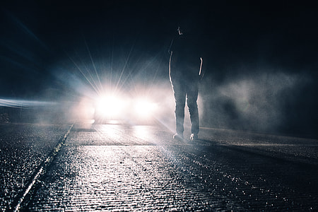 silhouette of man standing on asphalt road being lighted by a vehicle at night thumbnail
