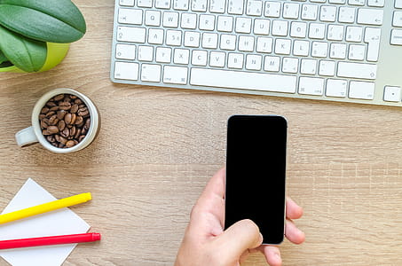 Person Holding Black Phone Near Apple Magic Keyboard on Brown Surface thumbnail