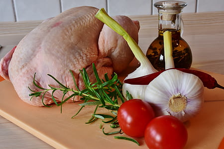 raw chicken beside oil cruet, garlic bulbs, chilli pepper, rosemary, and red tomatoes on beige chopping board closeup photo thumbnail