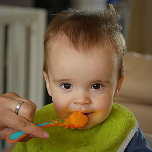 person's left hand holding food filled feeding spoon at baby's mouth thumbnail