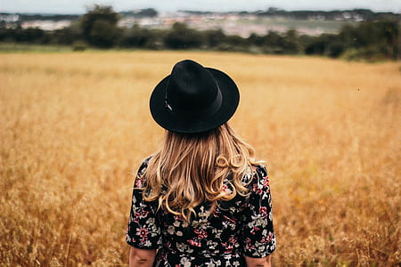 woman in black and multicolored floral short-sleeved top and black fedora hat standing on dried grass field thumbnail