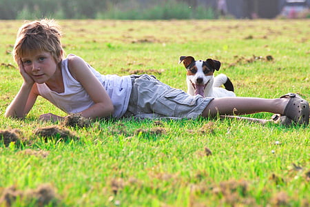 boy wearing white tank top lying on green grass beside short-coated white and brown dog during daytime thumbnail