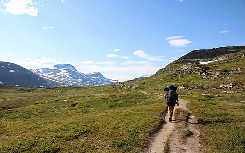 person with black hiking back walking on green field near mountains during daytime thumbnail