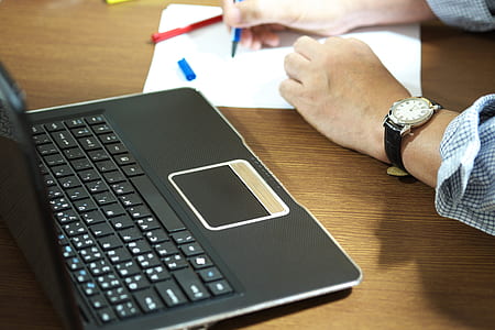 closeup photo of person holding blue pen writing on white paper beside black laptop computer thumbnail