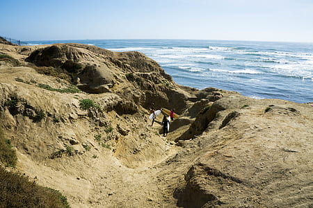 three people carrying surfboards on brown rock formation under blue sky thumbnail