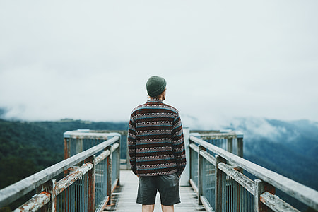 man wearing brown-and-grey striped sweater, blue denim jeans, and green cap standing in the middle of metal bridge overlooking view full of trees under cloudy sky during daytime thumbnail