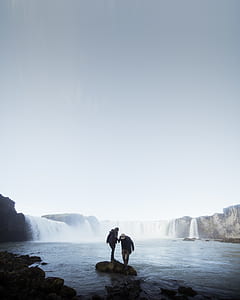 two person standing on rocks surrounded with water thumbnail