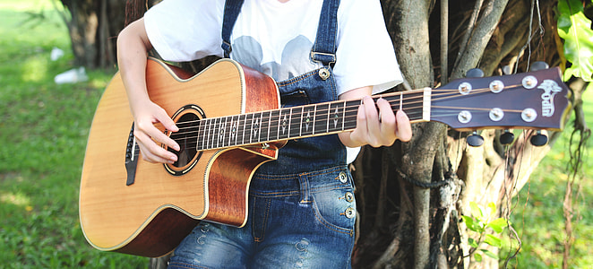 person wearing blue denim jumpshorts while playing guitar and leaning on brown tree thumbnail