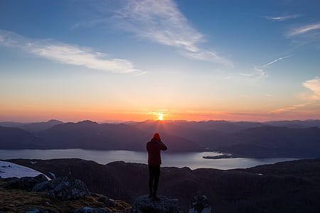 person standing on mountain peak facing body of water during sunrise thumbnail