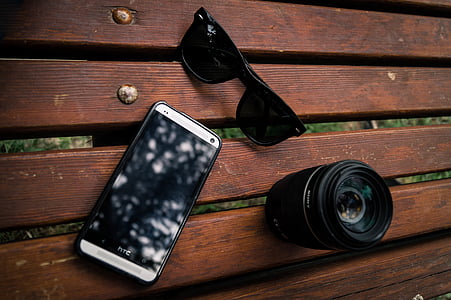 brown HTC smartphone, black wayfarer sunglasses and camera lens on brown wood platform thumbnail