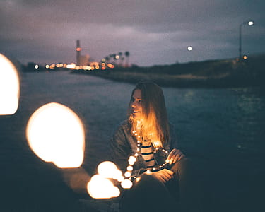 smiling woman with string lights on her neck near body of water at nighttime thumbnail