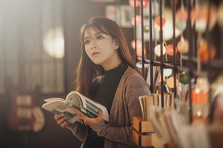 woman in brown cardigan holding book behind black metal book rack thumbnail
