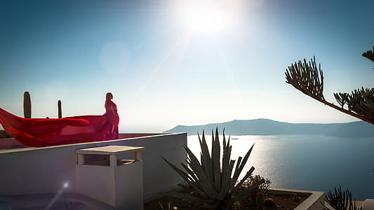 Woman Wearing Red Long Dress Standing at Rooftop of Building Facing Ocean thumbnail