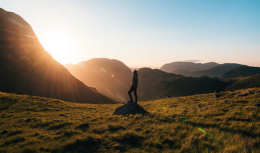 Silhouette Photography of Person Standing on Green Grass in Front of Mountains during Golden Hour thumbnail