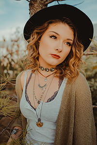 selective focus photography of woman wearing white Henley-neckline tank top, black hat and beige cardigan near tree thumbnail