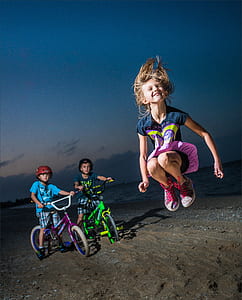 two boy's on bike and girl jumpig on seashore at night time thumbnail