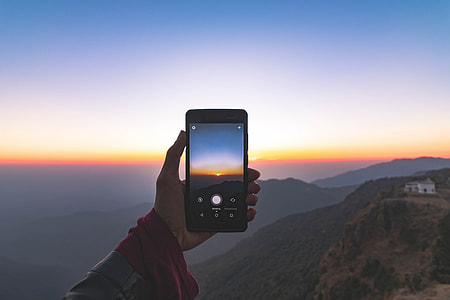 person taking photo of divided orange and blue skies over mountains near white house on mountain hill thumbnail