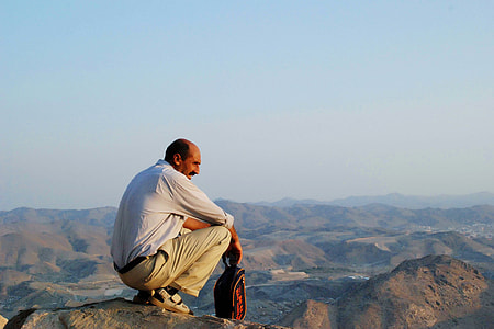 man wearing white dress shirt and khaki pants sitting on peak of mountain viewing hills at distance thumbnail