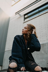 woman wearing blue denim jacket holding her hair beside white concrete wall during daytime thumbnail