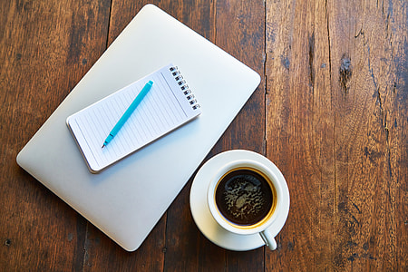 close-up photo of white cup, saucer, pocket notebook, and silver MacBook on brown wooden surface thumbnail