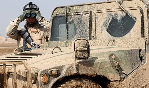 soldier wearing vest holding binoculars standing beside truck thumbnail