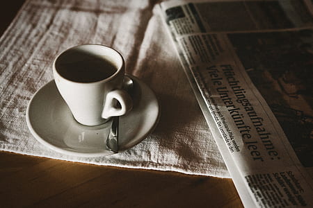 white ceramic teacup on top of white ceramic saucer plate with silver-colored spoon on it beside newspaper thumbnail