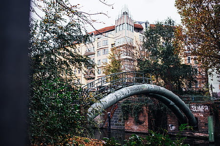 A curved metal pipeline with walkway railing over an urban river surrounded by buildings and trees thumbnail