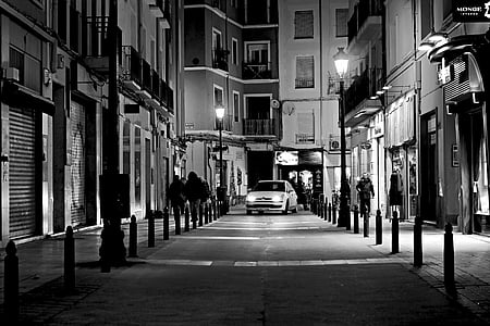 grayscale photo of white car passing on isle surrounded buildings thumbnail