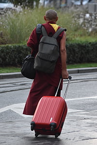 monk carrying bag while holding red luggage walking on road thumbnail