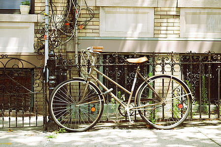 brown bicycle parked on sidewalk leaning on black steel fence thumbnail